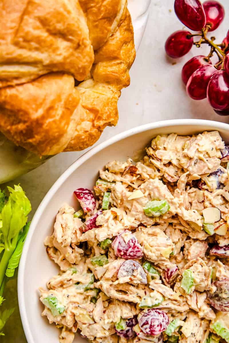 Traditional chicken salad in a bowl next to croissants and red grapes.
