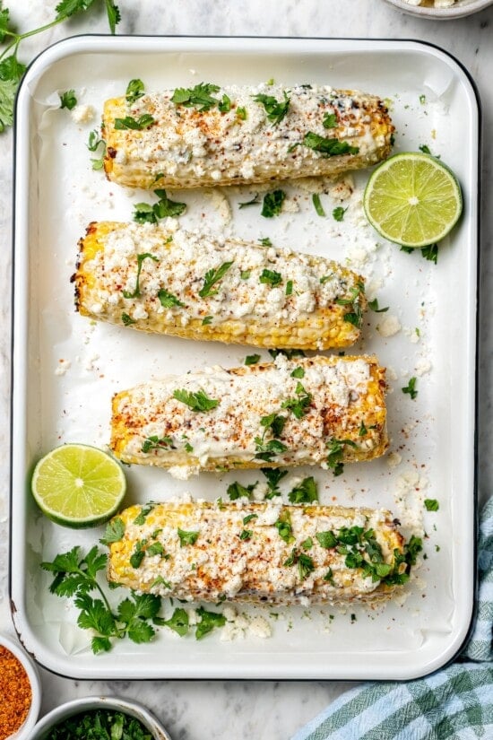 Overhead view of 4 ears of Mexican street corn in pan with lime halves and cilantro