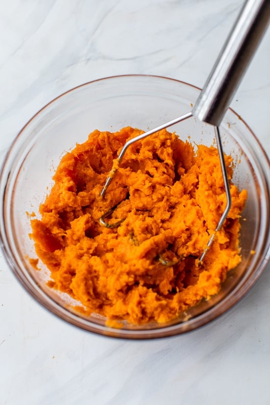 Potato masher mashing sweet potatoes in glass bowl.