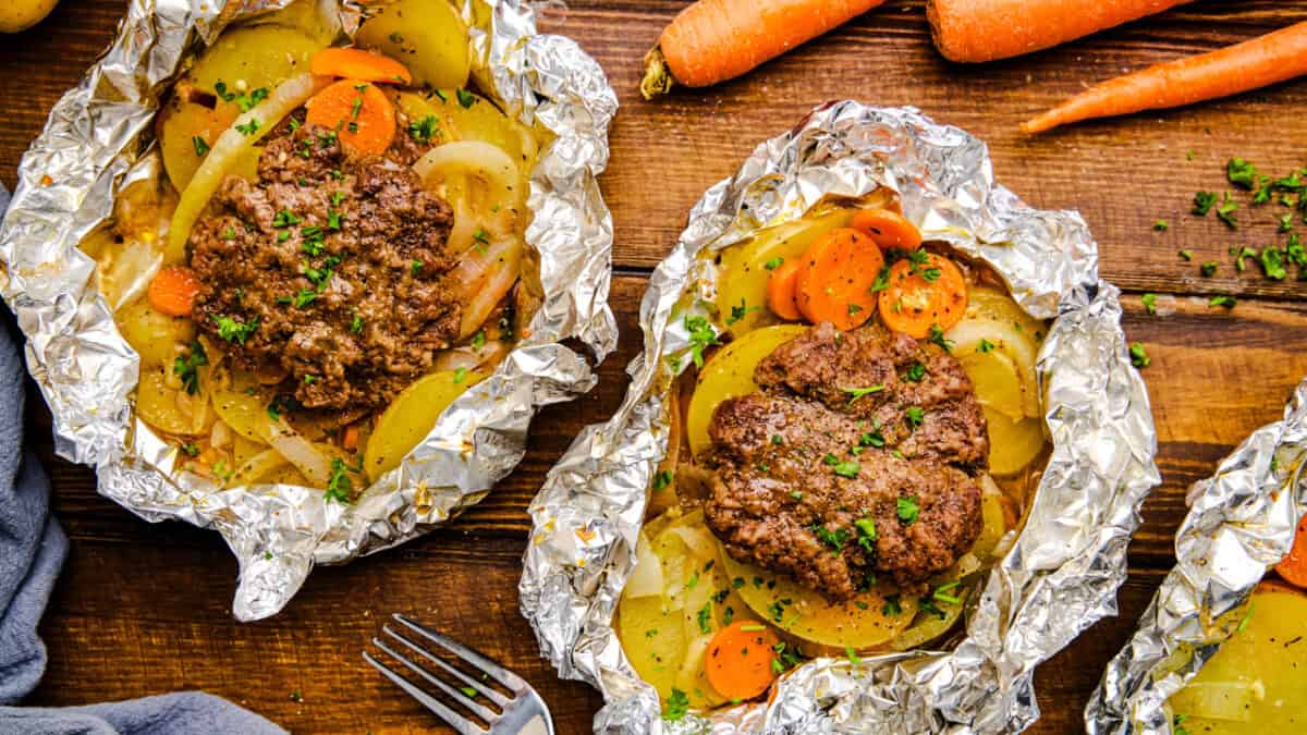 Open Hobo Foil Packet with burger, carrots, onions and potatoes on a wooden table.