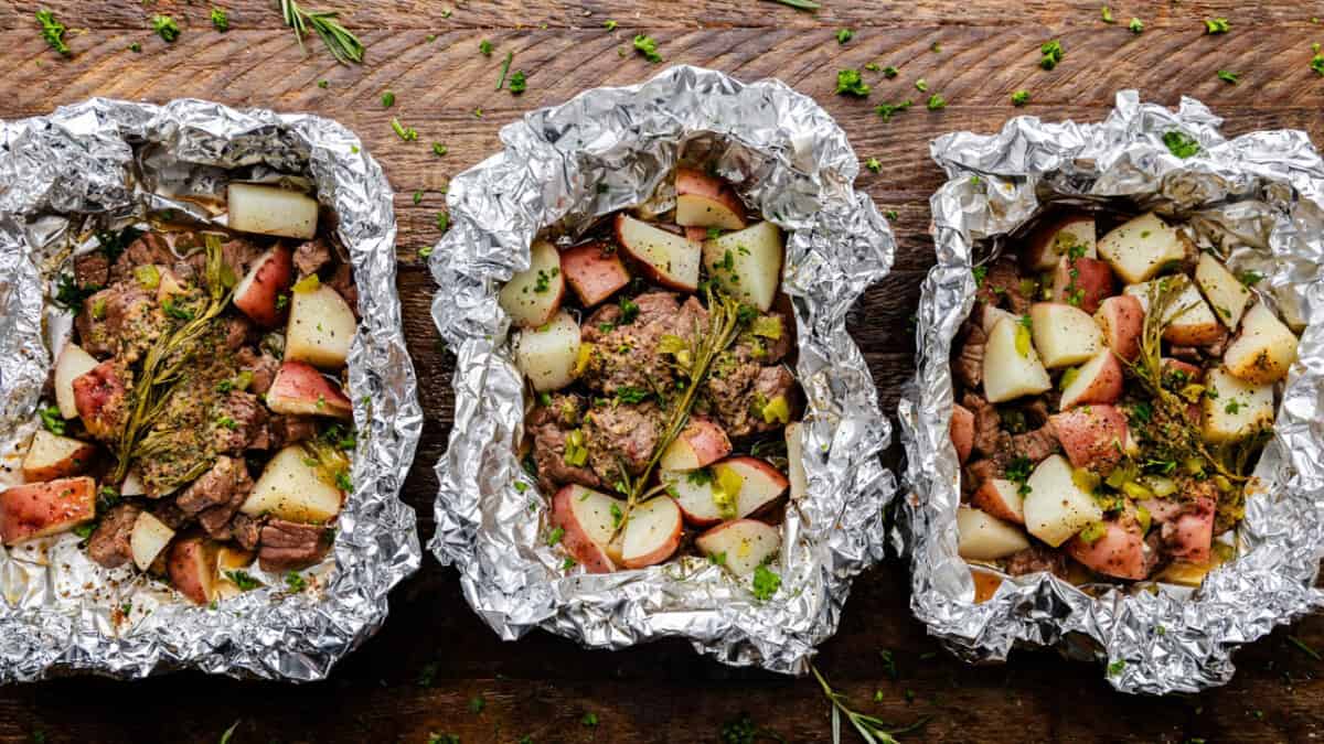 Three foil packets with cooked steak and potatoes.