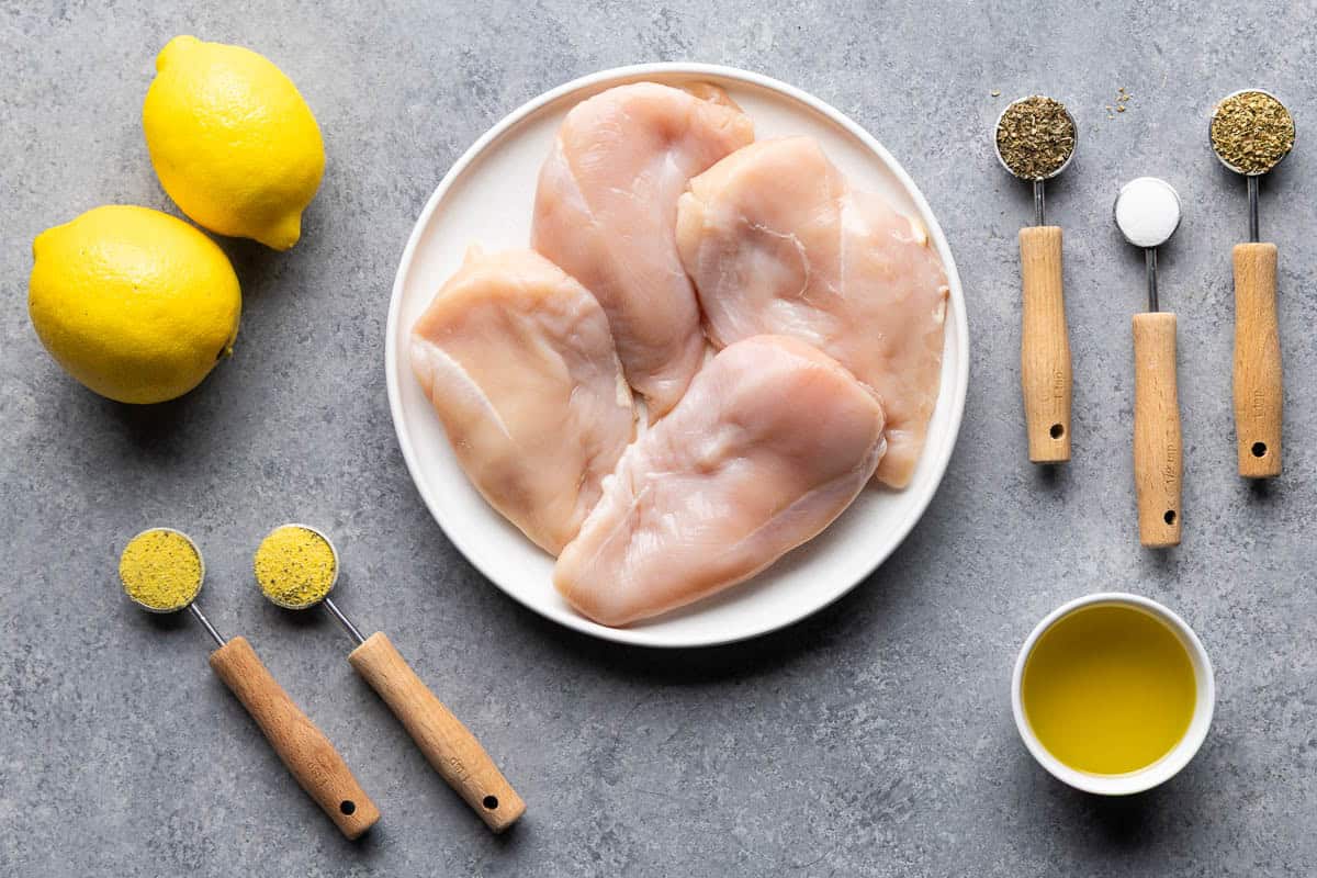 Ingredients for homemade lemon pepper chicken marinade arranged on a gray background, including raw chicken breasts, fresh lemons, olive oil, garlic seasoning, dried herbs, salt, and black pepper.