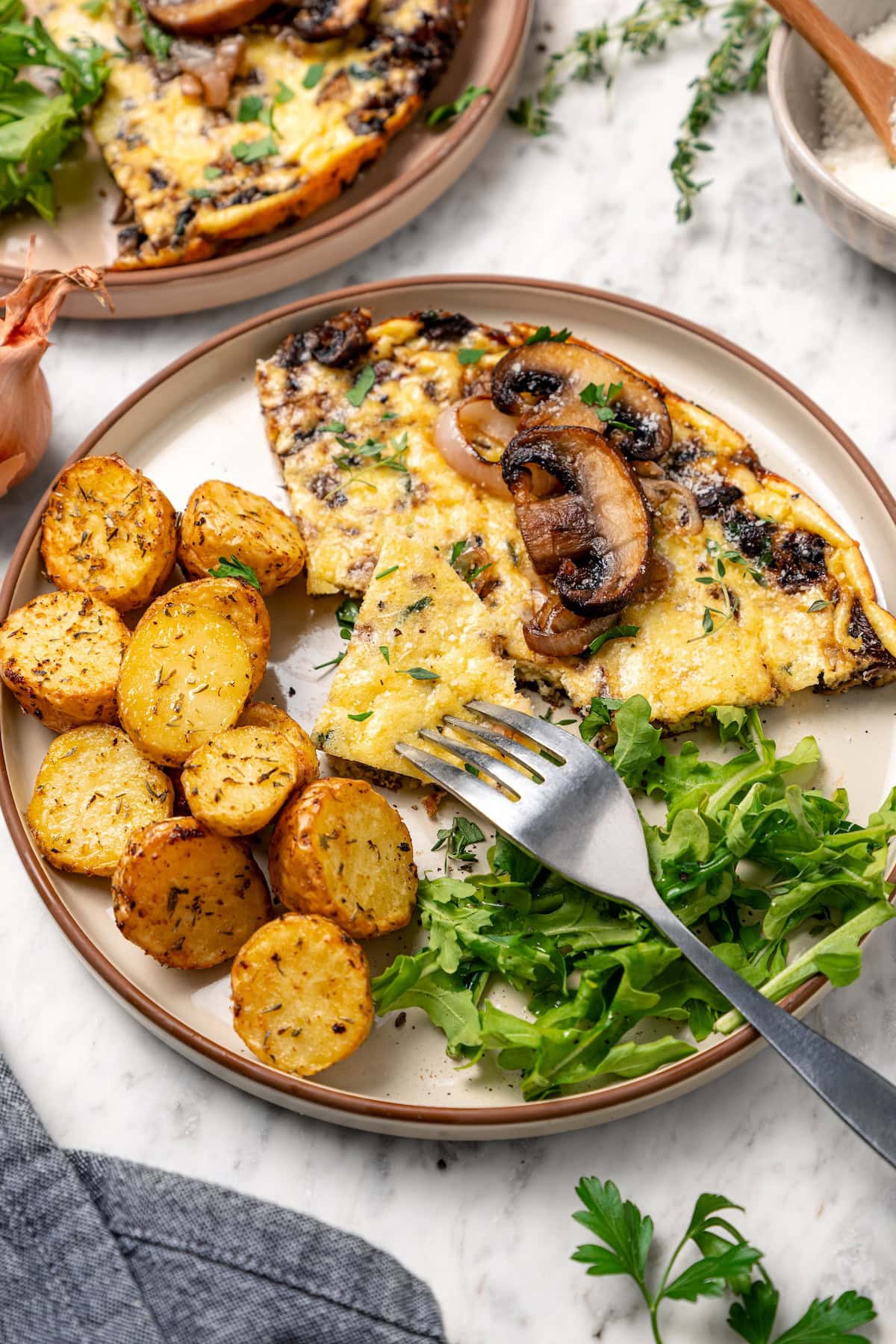 This aromatic mushroom frittata with egg whites, sautéed mushrooms, shallots, and cheese is an easy-to-make gourmet breakfast or brunch. A fork cutting into a slice of mushroom frittata served with crispy potatoes and fresh greens on a plate.
