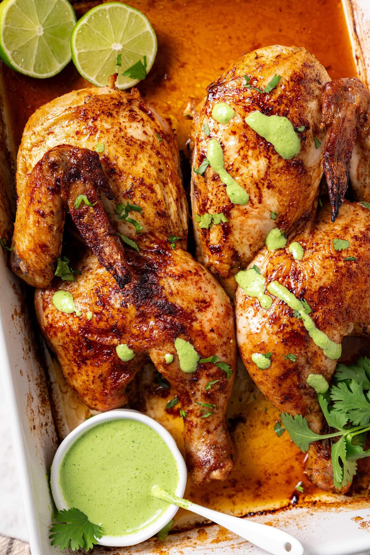This juicy, restaurant-style Peruvian chicken is marinated overnight and oven-roasted for golden, crispy skin. Served with spicy green sauce. Overhead view of crispy roasted Peruvian chicken in a baking dish with aji verde sauce and limes.
