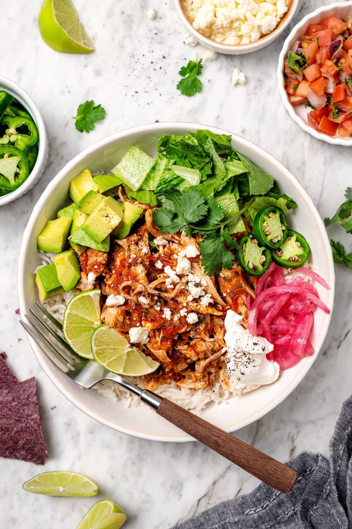 The easiest crockpot salsa chicken with juicy chicken thighs, jarred salsa, and simple spices. Great for meal prep, tacos, and rice bowls! Overhead view of a salsa chicken rice bowl garnished with avocado, pickled onions, jalapeño, lime wedges, and more toppings.
