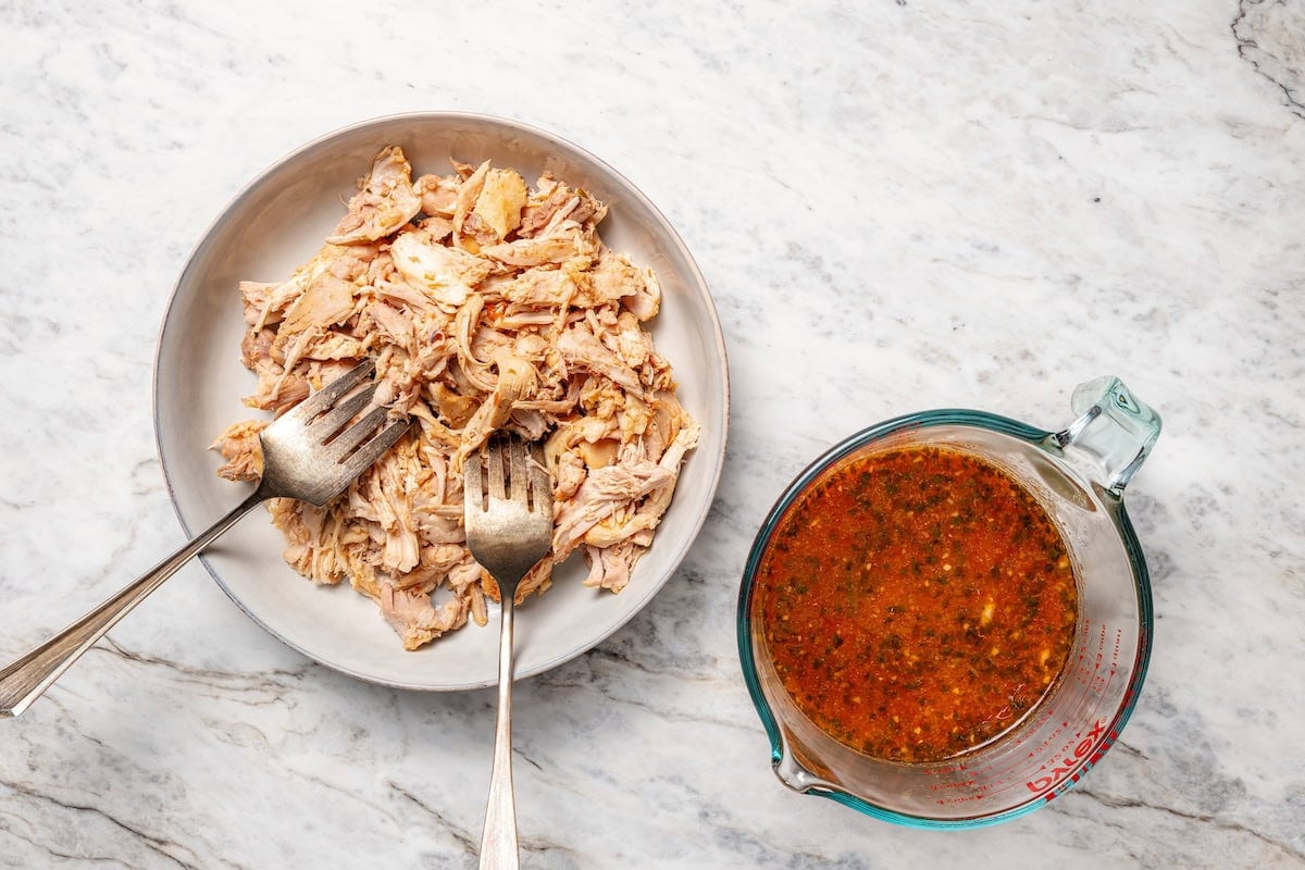 The easiest crockpot salsa chicken with juicy chicken thighs, jarred salsa, and simple spices. Great for meal prep, tacos, and rice bowls! Two forks shredding chicken on a plate next to a cup of cooking liquid.