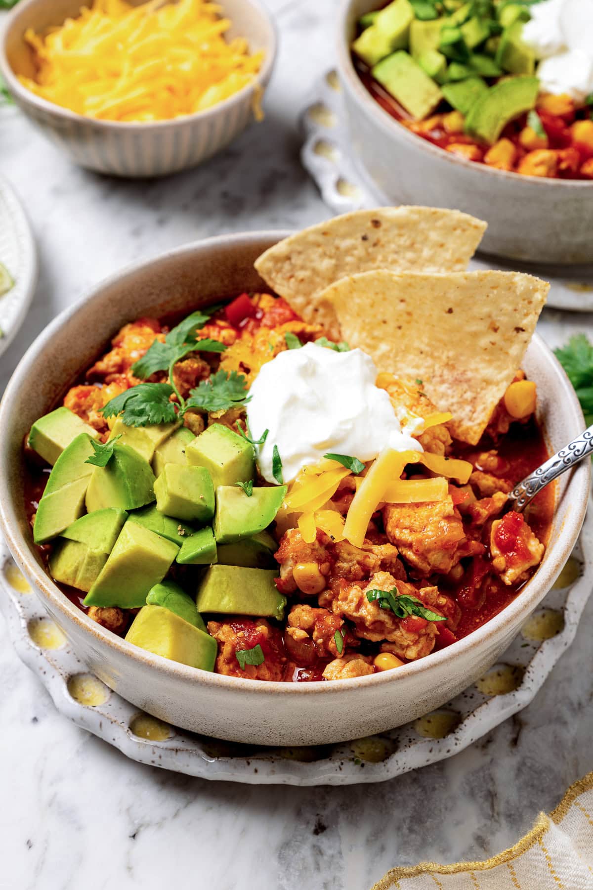 This crock pot turkey chili recipe is easy to make and family-friendly, with ground turkey, corn, bell pepper, tomatoes, and mild spices. A bowl of turkey chili garnished with sour cream, diced avocado, tortilla chips, and cilantro.